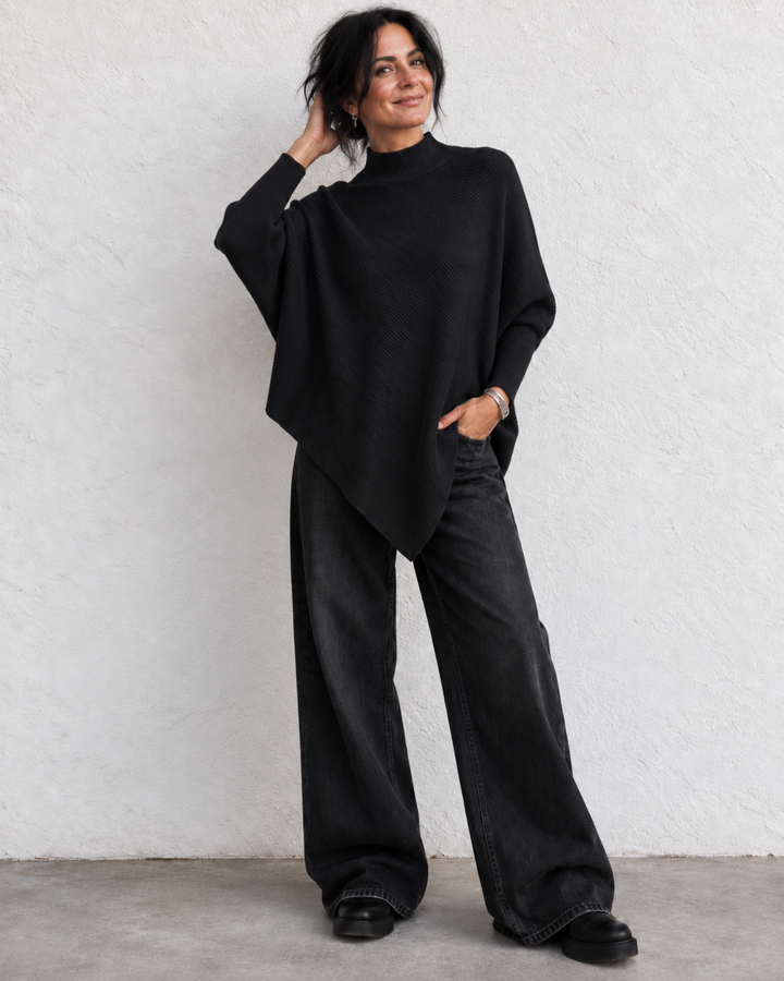A woman modeling a black long-sleeve knit poncho top with wide-leg charcoal gray jeans, standing against a textured white wall.