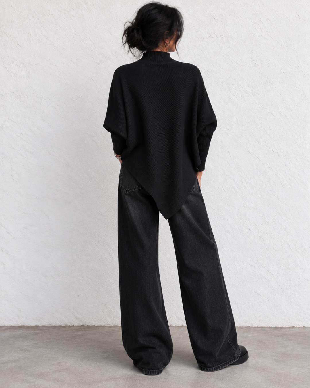 A person shown from the back wearing a loose black long-sleeve top and wide-leg black jeans, standing against a white textured wall on a light gray floor.