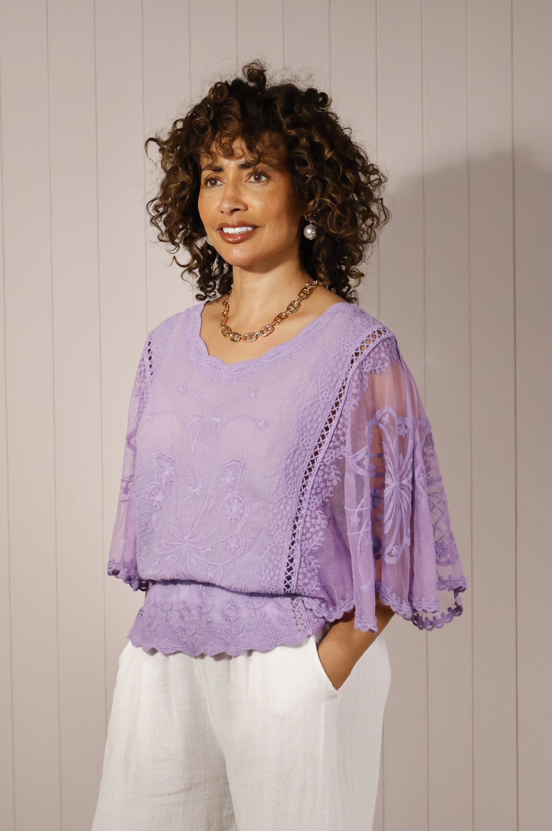 Woman wearing a purple lace top and white pants against a beige wall.