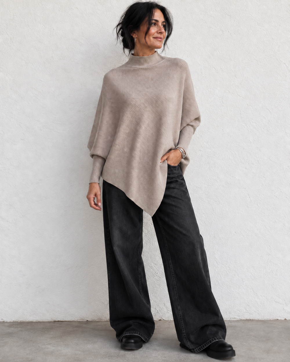 A woman wearing a taupe knit poncho sweater and wide-leg black jeans, standing against a textured white wall.