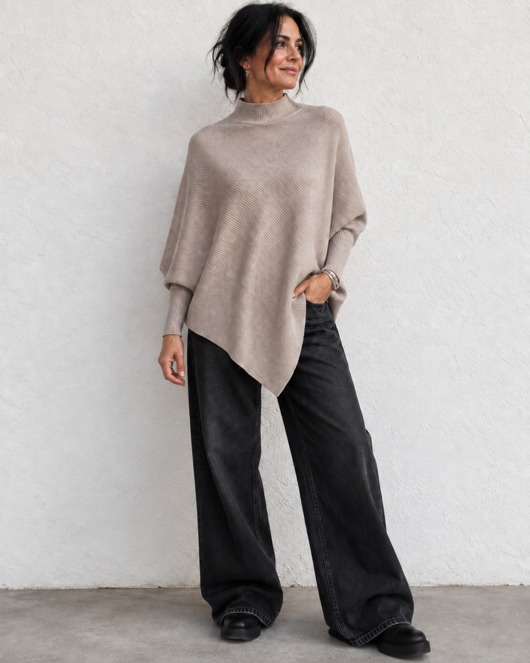 A woman wearing a taupe knit poncho sweater and wide-leg black jeans, standing against a textured white wall.