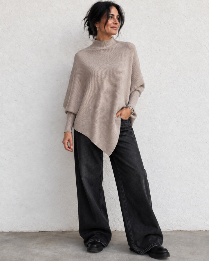 A woman wearing a taupe knit poncho sweater and wide-leg black jeans, standing against a textured white wall.
